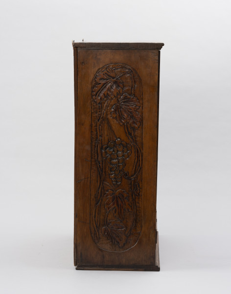 Szafka wisząca - ujęcie z lewego boku; ﻿Bok drewnianej, prostopadłościennej, wiszącej szafki. Ściana szafki zdobiona wzorem z kiści winogron.