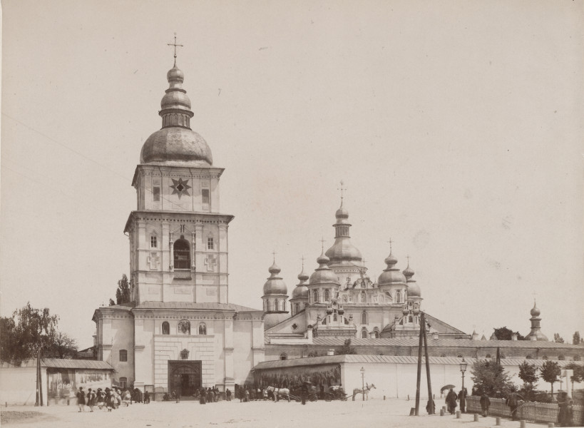 zbliżenie na zdjęcie z klasztorem. Po lewej dzwonnica nadbramna, przed którą grupki ludzi. Po prawej mur dekorowany malowidłami przedstawiającymi sceny religijne, w głębi sobór św. Michała