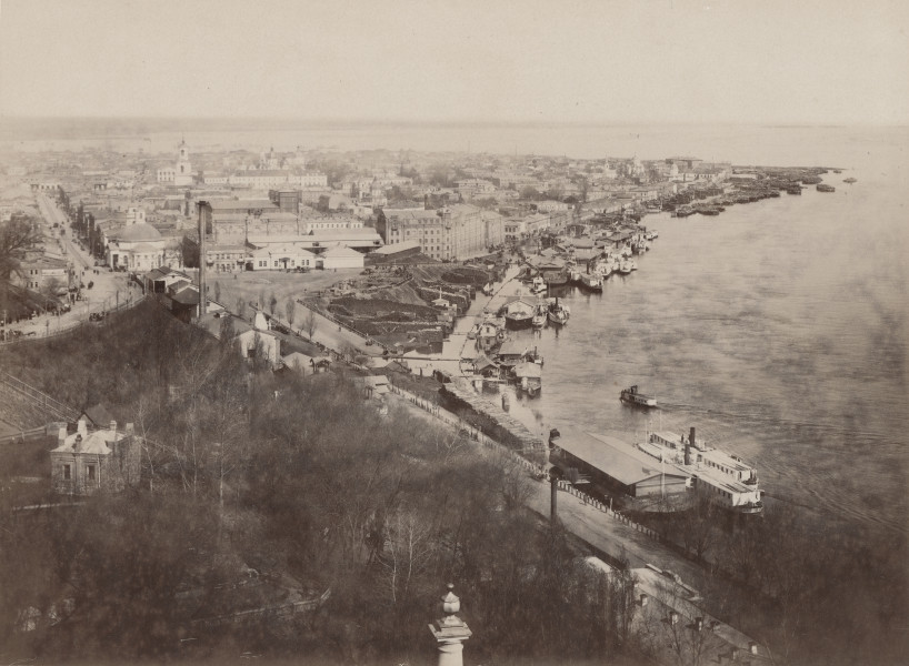 zbliżenie na zdjęcie przedstawiające widok miasta i nabrzeże rzeki z lotu ptaka. Na pierwszym planie bezlistne drzewa, po prawej barki na Dnieprze, po lewej zabudowa Kijowa