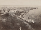zbliżenie na zdjęcie przedstawiające widok miasta i nabrzeże rzeki z lotu ptaka. Na pierwszym planie bezlistne drzewa, po prawej barki na Dnieprze, po lewej zabudowa Kijowa