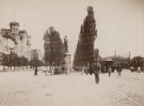 zbliżenie na zdjęcie z pomnikiem widzianym od prawej strony. W centrum fotografii monument, po prawej przystanek tramwajowy i pasażerowie, po lewej kamienice Kijowa