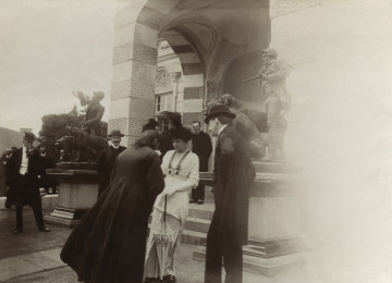 Na zdjęciu widoczne są roóżne osoby. Na pierwszym planie widoczny jest mężczyzna w marynarce i meloniku, a obok niego stoi kobieta w białej sukni i kapeluszu.