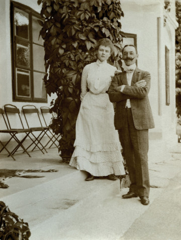 Na zdjęciu znajduje się kobieta w jasnej sukni, obok niej mężczyzna, na schodach przy pałacu.