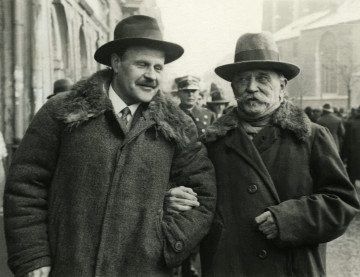 Na zdjęciu znajduje się dwóch mężczyzn w kapeluszach, idących pod ręke przez Krakowski Rynek.