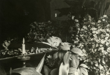 Fotografia pośmiertna; mężczyzna leżący w trumnie otoczonej kwiatami.