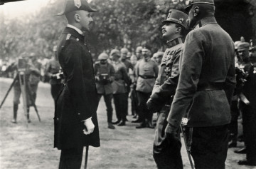 Na zdjęciu znajduje się trzech mężczyzn w mundurach na pierwszym planie, w tle widać innych mężczyzn w mundurach oraz fotografów.