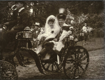 Na zdjęciu znajduje się para w strojach ślubnych jadąca powozem prowadzonym przez dwóch powożących mężczyzn.