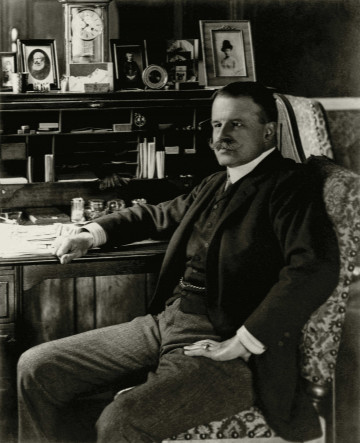 Zdjęcie przedstawia mężczyznę z wąsami, ubranego w garnitur, siedzącego przy biurku. 