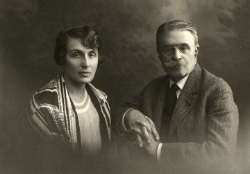 Na zdjeciu znajduje się kobieta i mężczyzna. Kobieta o ciemnych włosach, po lewej stronie; mężczyzna z wąsami, gładko zaczesanymi na bok włosami; w garniturze, siedzący po prawej stronie