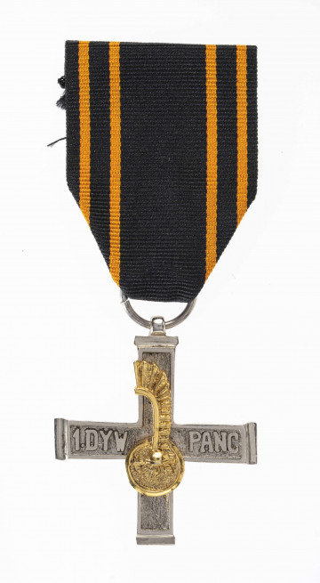 Odznaką Krzyża 1 Dywizji Pancernej jest krzyż bity w białym metalu o wymiarach 4,1 x 4,0 cm i szerokości ramion 0,7 cm. Powierzchnia krzyża z obydwu stron chropowata, obramowana wokół gładką ramką  szer. 0,1 cm. Na poziomych ramionach wypukły napis: 1 DYW. PANC. Na ramionach pionowych nałożona złocona miniaturka odznaki rozpoznawczej dywizji - szyszak na tle okrągłej tarczy ze skrzydłem husarskim. Krzyż noszony jest na czarnej wstążce szerokości 3,8 cm z 4 pomarańczowymi prążkami biegnącymi przy jej krawędziach.