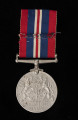 Medal Obrony - ujęcie z tyłu; Na rewersie: korona królewska spoczywająca na pniu dębu z liśćmi i owocami; po obydwu stronach korony i pnia stojące na tylnych łapach lew i lwica; nad grzbietami lwów daty: 1939 (nad lwem) i 1945 (nad lwicą); u dołu napis majuskułą: THE DEFENCE / MEDAL. Przy medalu nieprawidłowa wstążka (biało-granatowo-czerwona) od medalu 