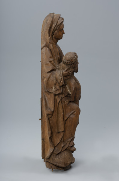 pieta - ujęcie z lewej; Rzeźba o pionowej kompozycji przedstawia dwufigurową grupę: Marię trzymającą martwe ciało Chrystusa. Sylwetka Marii wsparta jest na kamiennym zboczu porośniętym darnią widocznym po bokach i poniżej postaci. Głowę okrywa długi marszczony welon. Obszerny płaszcz nałożony na suknię o długich szerokich rękawach zagina się tworząc głębokie, światłocieniowe fałdowania i szerokie płaszczyzny na krawędziach. Ciało Chrystusa o nieco pomniejszonych proporcjach oddane jest naturalistycznie. Wsparte na kolanie matki osuwa się diagonalnie, prawa dłoń podtrzymuje głowę syna.  Wysuniętym do przodu i ustawionym nieco wyżej swym prawym kolanem podtrzymuje osuwające się diagonalnie martwe ciało Chrystusa o pomniejszonych proporcjach.