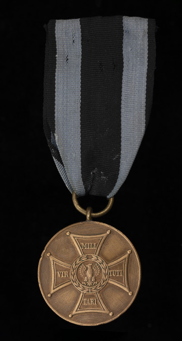 Medal Zasłużonym na Polu Chwały - ujęcie z przodu; Awers: na gładkim tle wizerunek Orderu Virtuti Militari; orzeł pośrodku krzyża bez korony. Medal zawieszony jest do czarnej wstążki z dwoma ciemnoniebieskim paskami.