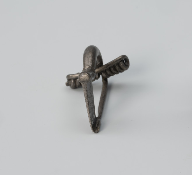 Fibula z wysoką pochewką - ujęcie z tyłu; Srebrna, niezdobiona zapinka z wysoką pochewką o prostej konstrukcji. Kabłąk ma postać masywnego drutu.