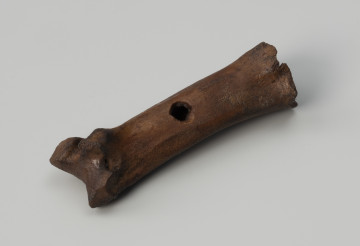 hetka - Ujęcie ze skosu z lewej; Hetka o zachowanej formie krótkiej kości śródstopia zwierzęcia z otworem przelotowym w środkowej części.
