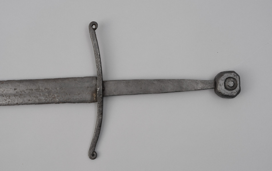 miecz typu XIIIa H2 1b (Oakeshott) - detal; Fragment miecza. Głownia o dł. 88,1 cm i szer. 4,9 cm, prosta, jednosieczna ze zbroczem, lekko zbieżna ku sztychowi. Sztych kończysty. Trzpień głowni lekko zwężający się ku końcowi. Jelec krzyżowy, lekko wygięty ku głowni, z zawiniętymi ku głowicy końcami ramion. Głowica nieregularna, ośmioboczna, z rytym dużym kołem na płaszczyznach. Znaki i zdobienia: Na głowni, złotym metalem, nieczytelny znak, dwóch okręgów, w środku których, znajdował się znak krzyża kawalerskiego, obecnie słabo czytelny.