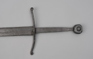 miecz typu XIIIa H2 1b (Oakeshott) - detal; Fragment miecza. Głownia o dł. 88,1 cm i szer. 4,9 cm, prosta, jednosieczna ze zbroczem, lekko zbieżna ku sztychowi. Sztych kończysty. Trzpień głowni lekko zwężający się ku końcowi. Jelec krzyżowy, lekko wygięty ku głowni, z zawiniętymi ku głowicy końcami ramion. Głowica nieregularna, ośmioboczna, z rytym dużym kołem na płaszczyznach. Znaki i zdobienia: Na głowni, złotym metalem, nieczytelny znak, dwóch okręgów, w środku których, znajdował się znak krzyża kawalerskiego, obecnie słabo czytelny.