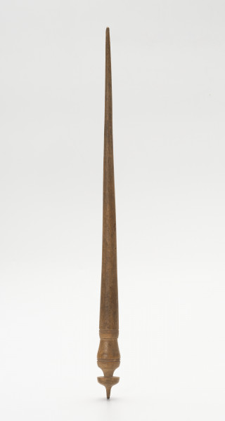wrzeciono - Ujęcie z przodu; Wrzeciono wykonane z drewna w kształcie iglicy. Częściowo profilowane, w dolnej części zakończone pierścieniem przechodzącym w krótki szpic i dookolnie ryte.