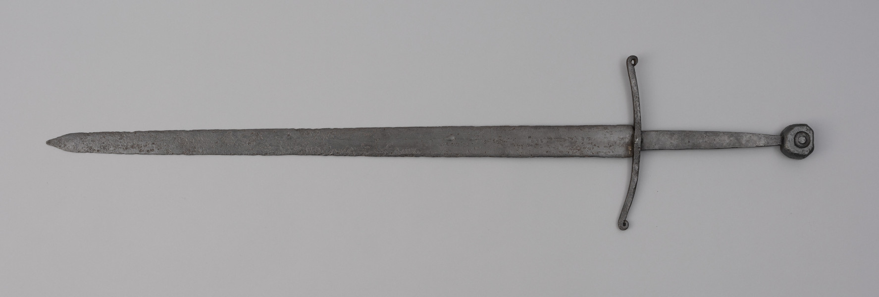miecz typu XIIIa H2 1b (Oakeshott) - ujęcie z tyłu; Miecz w typie: XIIIa, H2, 1b. Głownia o dł. 88,1 cm i szer. 4,9 cm, prosta, jednosieczna ze zbroczem, lekko zbieżna ku sztychowi. Sztych kończysty. Trzpień głowni lekko zwężający się ku końcowi. Jelec krzyżowy, lekko wygięty ku głowni, z zawiniętymi ku głowicy końcami ramion. Głowica nieregularna, ośmioboczna, z rytym dużym kołem na płaszczyznach. Znaki i zdobienia: