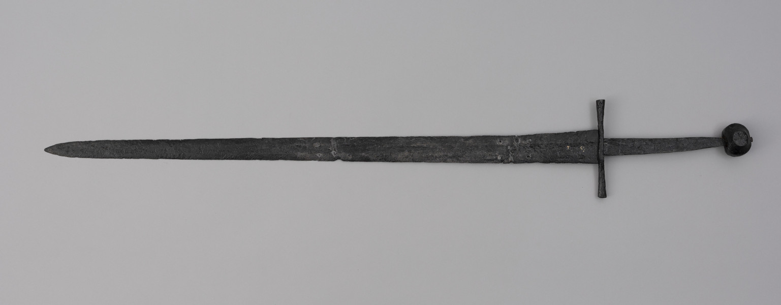 miecz typu XIVa J2 (Oakeschott) - ujęcie z tyłu; Miecz długi półtoraręczny. Głownia długości 95 cm i szer. u nasady 5,6 cm, prosta dwusieczna zbieżna ku sztychowi, łączona wtórnie w dwóch miejscach. Zbrocze długie, sięga 3/4 długości. Pierwotnie po obu stronach głowni znajdował się napis ryty, wypełniony żółtym metalem w postaci krzyżyka oraz trzech liter, z których pierwsza jest nieczytelna, ale przypomina L lub T; druga, środkowa to D; trzecia litera to połączenie I i H. Trzpień rękojeści zwężający się ku końcowi. Jelec krzyżowy o długości 16,7 cm, z rozszerzającymi się ramionami. Głowica kolista, fazowana, dwubocznie spłaszczona o wys. 4,3 cm i szer. 5,6 cm. Powierzchnia z wżerami pokorozyjnymi. Typ wg Oakeschotta: XIVa J2 (ewentualnie XIIIa).