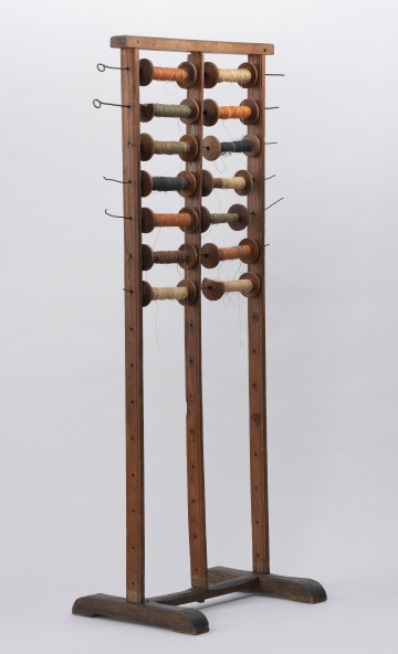 Szpularz - ujęcie ze skosu z lewej; Szpularz zbudowany z trzech pionowo, równolegle względem siebie ustawionych listew, u góry połączonych krótszą i grubszą listwą poprzeczną, u dołu osadzonych w mającej kształt przypominający literę H podstawie. W listwach pionowych 14 rzędów wywierconych niewielkich otworów, w których umieszczone są metalowe pręty z osadzonymi na nich (po dwie) drewnianymi szpulami z nawiniętymi na nie kolorowymi nićmi. Ilość szpul: 14