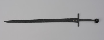 miecz typu XIVa J2 (Oakeschott) - ujęcie z przodu; Miecz długi półtoraręczny. Głownia długości 95 cm i szer. u nasady 5,6 cm, prosta dwusieczna zbieżna ku sztychowi, łączona wtórnie w dwóch miejscach. Zbrocze długie, sięga 3/4 długości. Pierwotnie po obu stronach głowni znajdował się napis ryty, wypełniony żółtym metalem w postaci krzyżyka oraz trzech liter, z których pierwsza jest nieczytelna, ale przypomina L lub T; druga, środkowa to D; trzecia litera to połączenie I i H. Trzpień rękojeści zwężający się ku końcowi. Jelec krzyżowy o długości 16,7 cm, z rozszerzającymi się ramionami. Głowica kolista, fazowana, dwubocznie spłaszczona o wys. 4,3 cm i szer. 5,6 cm. Powierzchnia z wżerami pokorozyjnymi. Typ wg Oakeschotta: XIVa J2 (ewentualnie XIIIa).