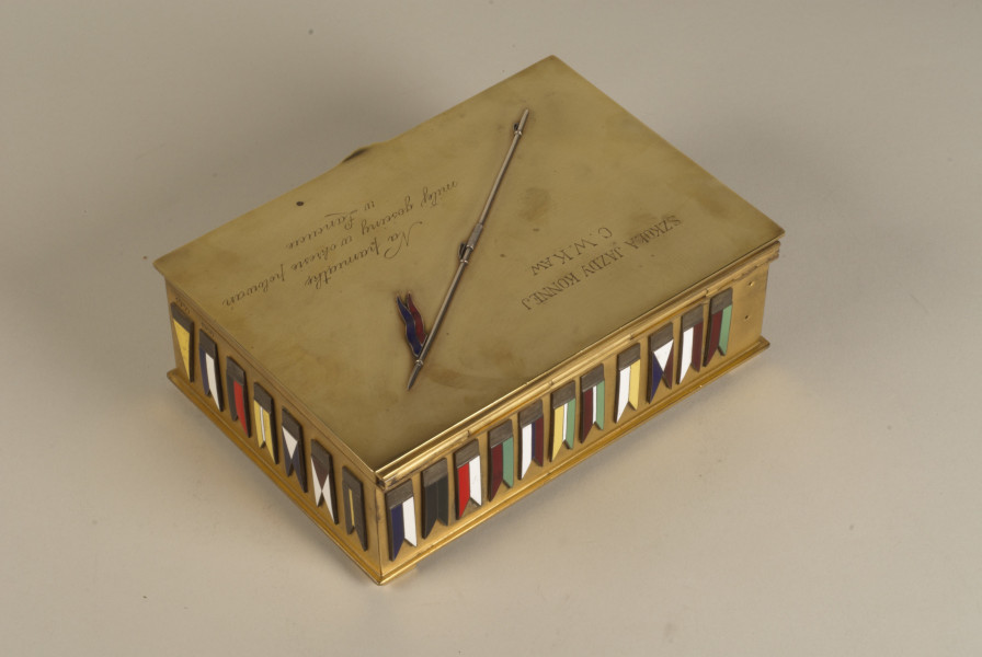 Pamiątkowa kasetka na cygara zdobiona grawerunkami i emaliowanymi proporcami kawaleryjskimi. Wykonana z mosiądzu (złoconego?), w kształcie prostopadłościanu z otwieranym na zawiasach wiekiem