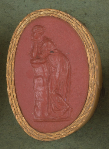 czerwona owalna gemma w grubym złotym obramowaniu; kobieta oparta w zamyśleniu o kamienny murek, podpiera głowę na ręce, ma włosy spięte w kok, jest ubrana w chiton i himation