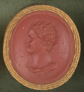 czerwona owalna gemma w grubym złotym obramowaniu; lewy profil młodego, uśmiechniętego mężczyzny w wieńcu z winorośli, pod szyją widoczny fragment szaty; za głową postaci ledwie widoczna sygnatura 