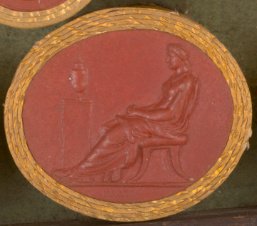 czerwona gemma, owalna pozioma, w grubym złotym otoku. Lewy profil siedzącej kobiety ze związanymi na karku włosami i złożonymi na kolanach rękoma. Przed nią, na postumencie - urna z prochami zmarłego męża, Germanika.