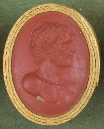 czerwona owalna gemma w grubym złotym obramowaniu; prawy profil mężczyzny o krótkich włosach i brodzie oraz o orlim nosie, wida umięśniony tors i ramiona
