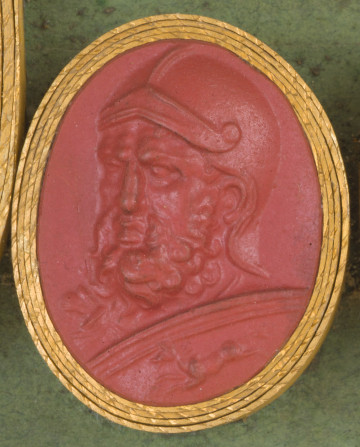 czerwona owalna gemma w grubym złotym obramowaniu; widoczna z półprofilu twarz wojownika z hełmem na głowie, z długą kręconą brodą i wąsami, poniżej widoczny fragment okrągłej tarczy, na której widać galopującego konia