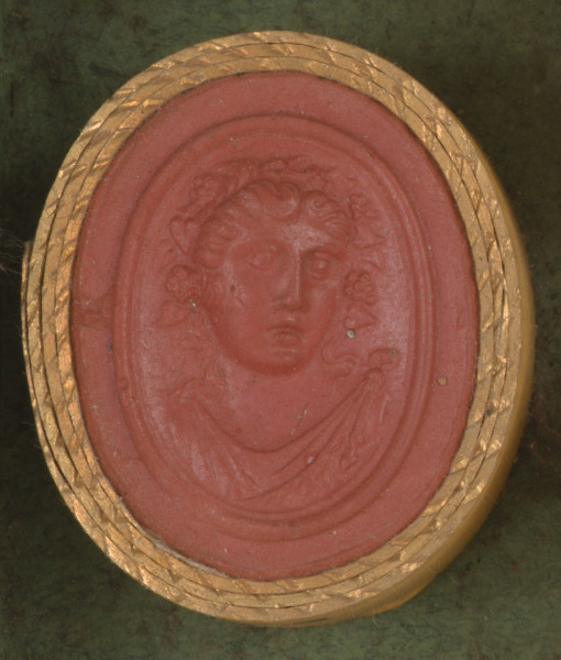 czerwona owalna gemma w grubym złotym obramowaniu; głowa postaci (prawdopodobnie kobiety) w wieńcu z winorośli, widzoczna na wprost, poniżej widać fragment szaty