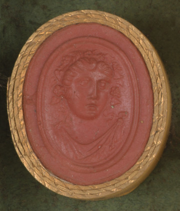 czerwona owalna gemma w grubym złotym obramowaniu; głowa postaci (prawdopodobnie kobiety) w wieńcu z winorośli, widzoczna na wprost, poniżej widać fragment szaty