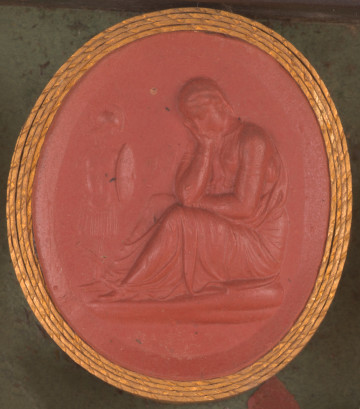czerwona gemma, owalna pionowa, w grubym złotym obramowaniu. Siedząca kobieta w długiej szacie, ukazana w prawym pólprofilu. Nogi zgiete w kolanach, głowa podparta na prawej dłoni. W tle po lewej zbroja rzymska z hełmem, tarczą i mieczem.