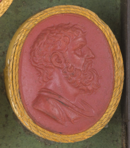 czerwona owalna gemma w grubym złotym obramowaniu; widoczne w prawego profilu popiersie mężczyzny w średnim wieku, z krótkimi kręconymi włosami, wąsami i brodą, poniżej wida fragment tuniki