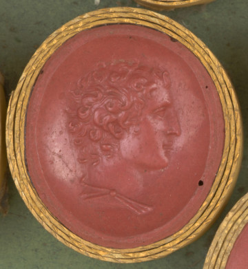 czerwona owalna gemma w grubym złotym obramowaniu, młody mężczyzna widoczny z prawego profilu, ma krótkie kręcone włosy, na ramieniu widoczna część tuniki z zapięciem