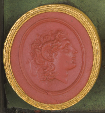 czerwona owalna gemma w grubym złotym obramowaniu; prawy profil głowy młodego mężczyzny z lekko kręconymi włosami i cienką opaską na włosach, bez zarostu