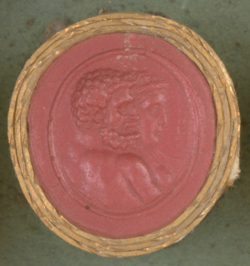 czerwona owalna gemma w grubym złotym obramowaniu, na pierwszym planie profil Herkulesa z kręconymi krótkimi włosami, brodą i wąsami, z nagim torsem. Na drugim planie profil kobiety z długimi włosami i diademem na głowie.