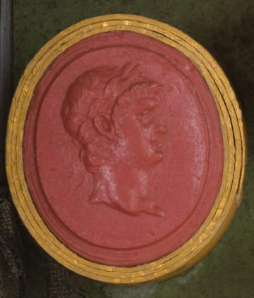 czerwona owalna gemma w grubym złotym obramowaniu; prawy profil głowy młodego mężczyzny z krótkimi włosami i wieńcem laurowym związanym z tyłu, ma ledwie widoczny bardzo krótki zarost. 