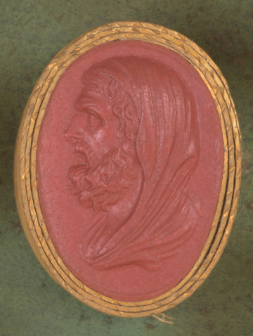 czerwona owalna gemma w grubym złotym obramowaniu; lewy profil popiersiamężczyzny w średnim wieku, z kręconymi włosami, bujną brodą i wąsami; mężczyzna ma kaptur na głowie