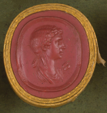 czerwona owalna gemma w grubym złotym obramowaniu; kobieta widoczna z prawego profilu, ma półdługie włosy upięte z tyłu, widoczny jest fragment szaty, za nią dwa kłosy zboża