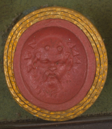 czerwona owalna gemma w grubym złotym obramowaniu; maska w kształcie twarzy starszego mężczyzny z prostą brodą i wąsami; mężczyzna jest łysy i ma małe rogi wyrastające z czoła i liście laurowe na głowie