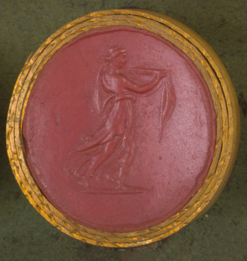czerwona okrągła gemma w grubym złotym obramowaniu; młoda kobieta widoczna z prawego profilu, ubrana w zwiewny peplos, niosąca w wyciągniętych przed siebie rękach misę
