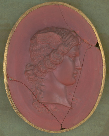 czerwony wycisk owalny ze złotym obramowaniem; prawy profil głowy kobiety z kręconymi włosami spiętymi z tyłu, z przodu na głowie widoczne małe węże, z boku głowy skrzydełka; gemma uszkodzona: widoczne pęknięcia i niewielkie ubytki po bokach