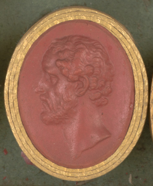 czerwona owalna gemma w grubym złotym obramowaniu, lewy profil mężczyzny w dojrzałym wieku, mężczyzna ma krótkie kręcone włosy, wąsy i krótką brodę