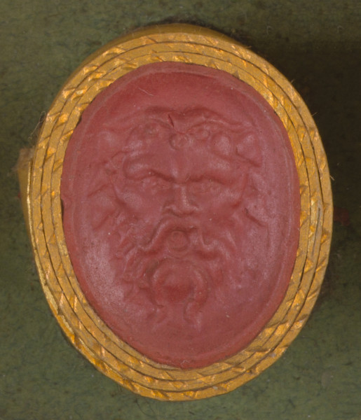 czerwona owalna gemma w grubym złotym obramowaniu; maska przedstawia głowę Sylena, Syle nma na głowie rogi, kręcone włosy, wąsy i brodę; ma groźny wyraz twarzy: zmarszczone brwi i otwarte usta.