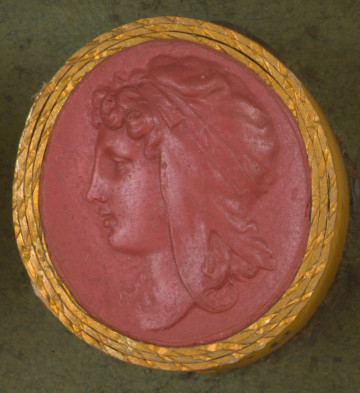 czerwona okrągła gemma w grubym złotym obramowaniu; głowa młodego mężczyzny z długimi kręconymi włosami widziana z lewego profilu; mężczyzna ma na głowie czapke frygijską