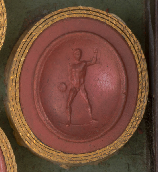 czerwona owalna gemma w grubym złotym obramowaniu; młody nagi mężczyzna stojący na wprost widza, w prawej ręce trzyma dysk lub kulę, lewą ma uniesioną do góry