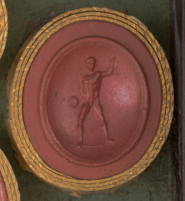 czerwona owalna gemma w grubym złotym obramowaniu; młody nagi mężczyzna stojący na wprost widza, w prawej ręce trzyma dysk lub kulę, lewą ma uniesioną do góry